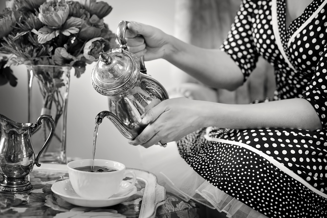 tea-party-1001654_1280.jpg A person in a polka dot dress pours tea from a teapot into a cup beside a vase of flowers on a table.