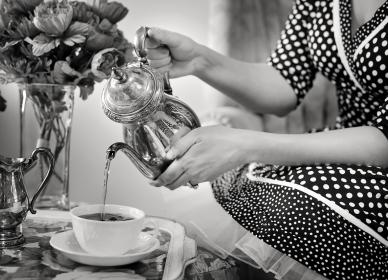 A person in a polka dot dress pours tea from a teapot into a cup beside a vase of flowers on a table.