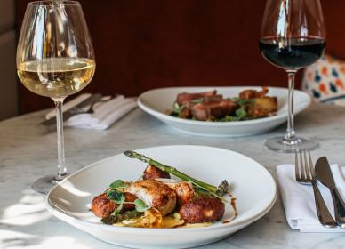 A plate of roasted chicken with vegetables and sauce, served with white wine; another plate with meat and red wine is in the background on a marble table.